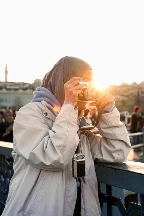 Free A woman takes a photo on a bridge with sunlight in the background, capturing a cityscape during sunset. Stock Photo