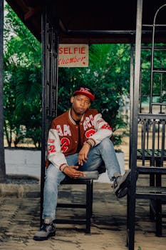 A young man sits casually in a selfie chamber, wearing a trendy jacket and cap.