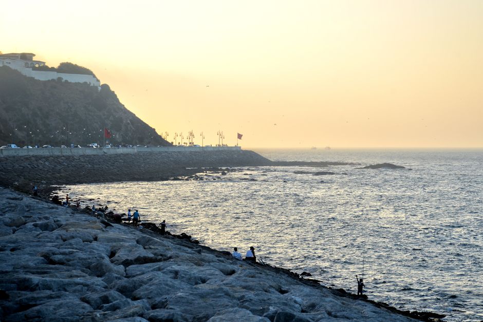 Peaceful coastal evening scene with people fishing, a rocky shoreline, and cliffs at sunset.