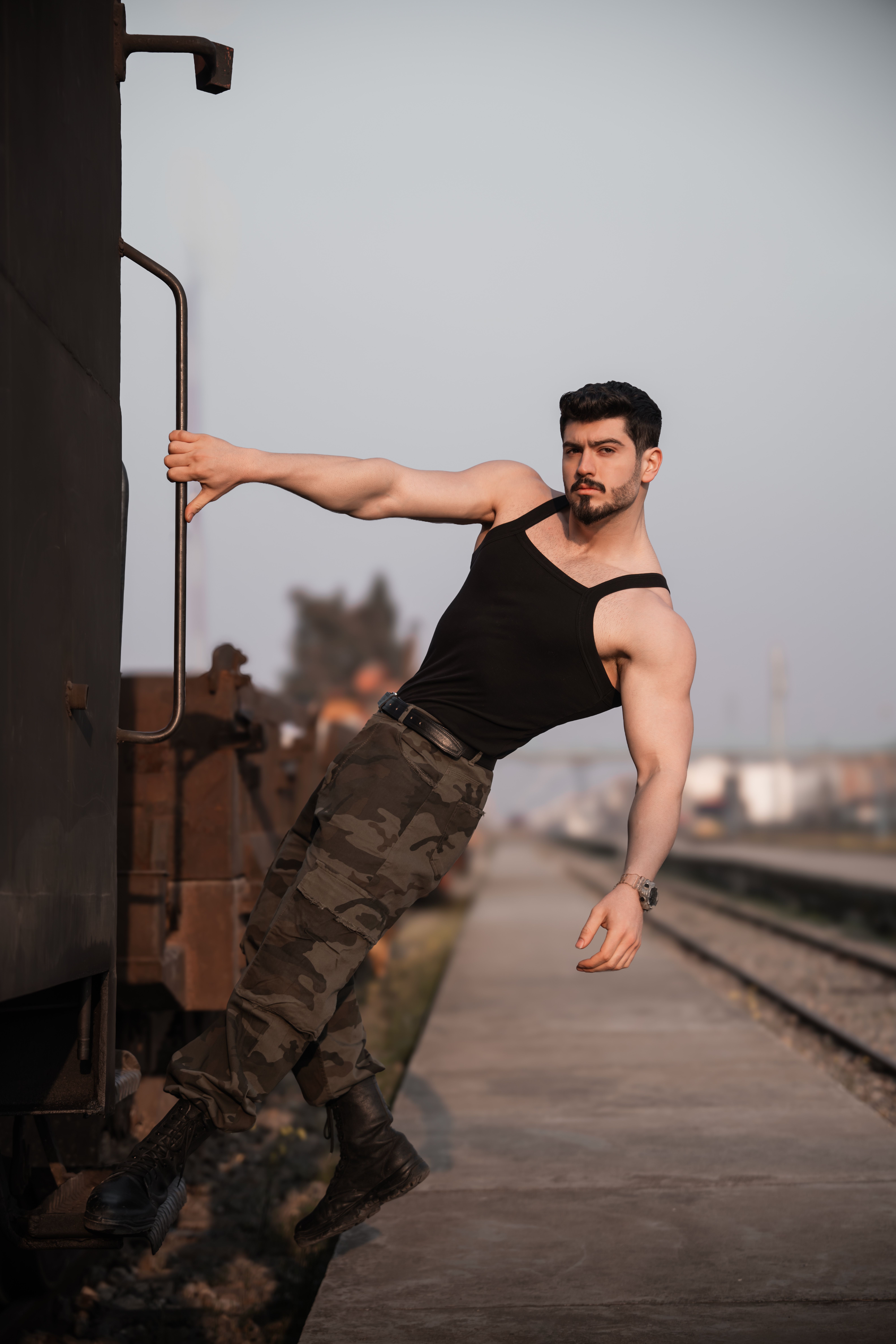 Muscular man in black tank top and camo pants posing by a train at sunset for a strong outdoor portrait.