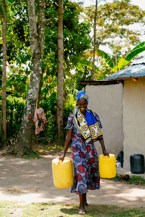 Free Smiling African woman carrying yellow water containers outdoors near a rural home under bright day. Stock Photo