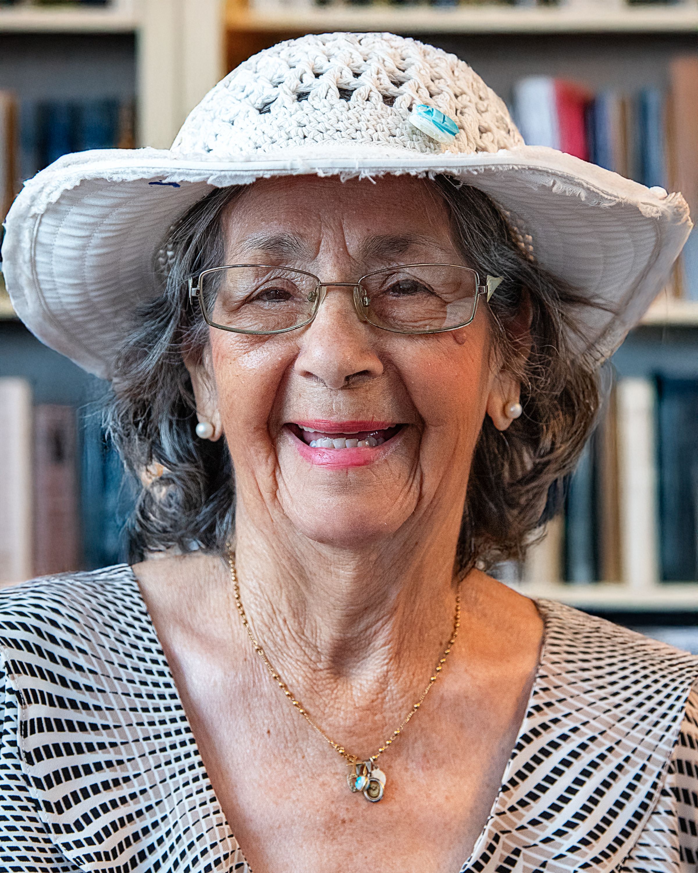 Elderly woman smiling in a library, wearing a white hat and glasses, exuding warmth and joy.