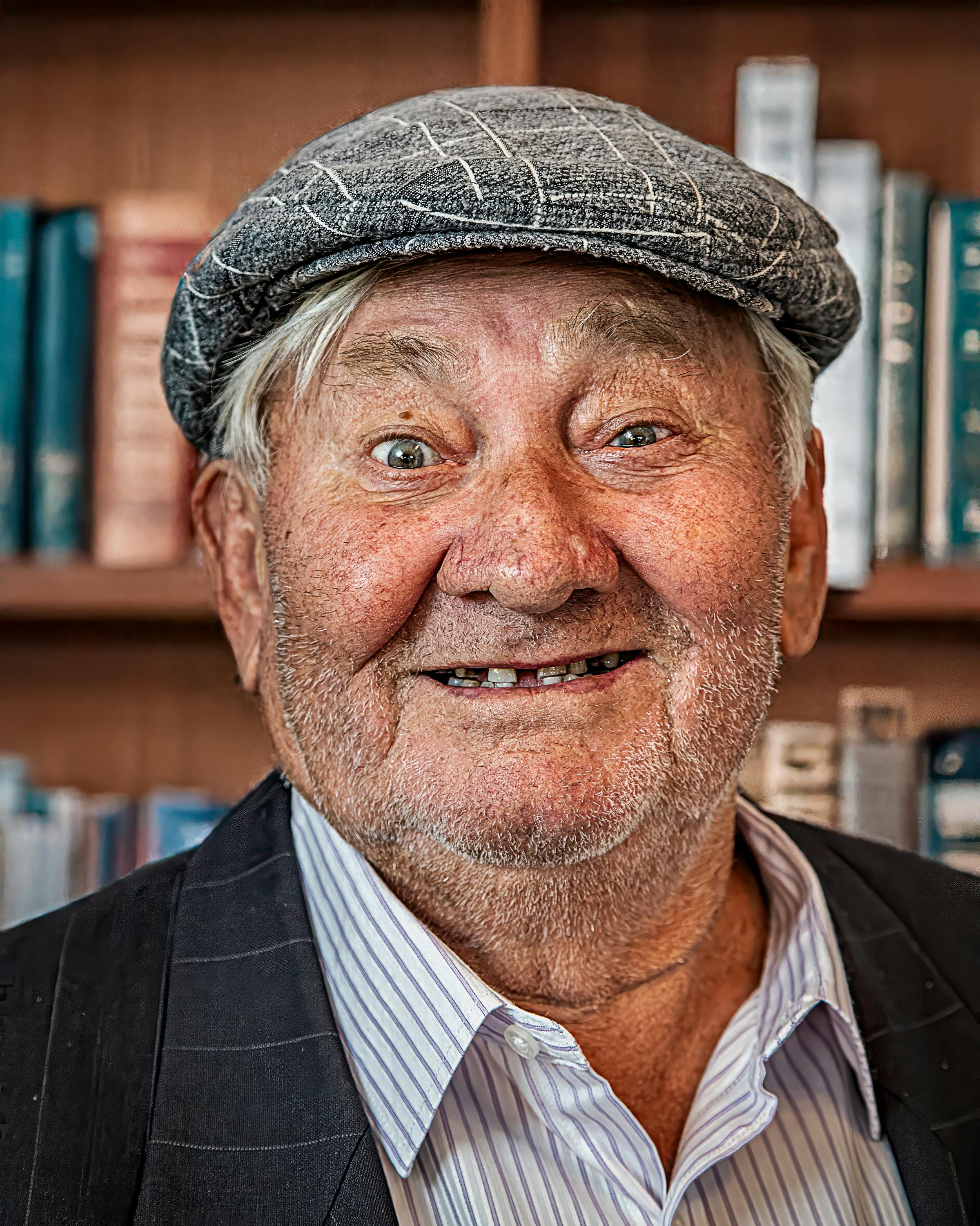 Portrait of a cheerful elderly man wearing a cap, smiling against a bookshelf backdrop.