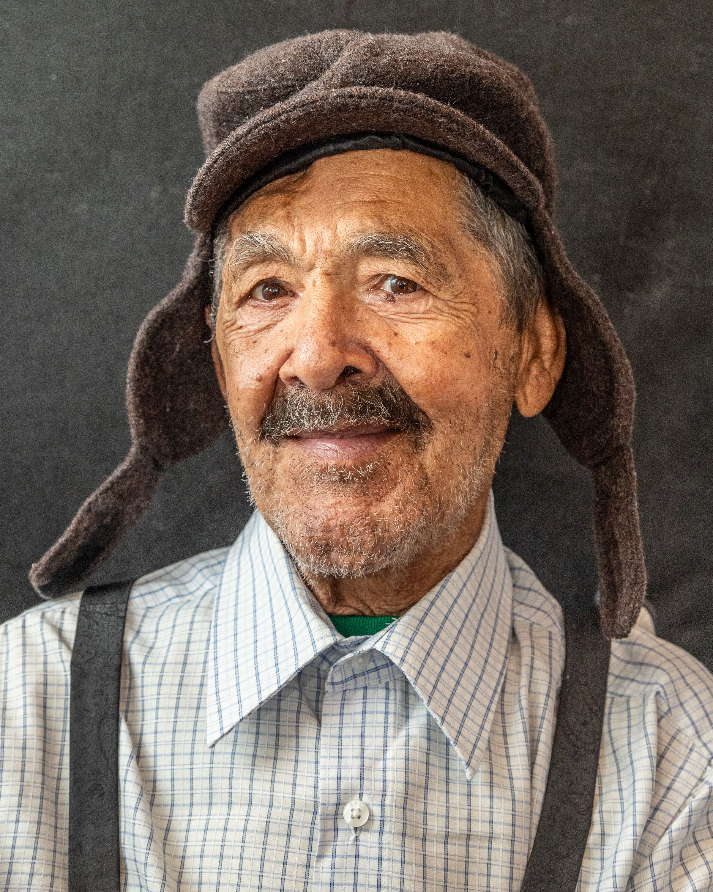Elderly man in plaid shirt and warm hat, smiling warmly at the camera.