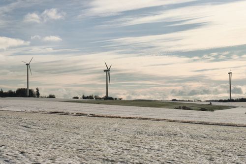 Free Serene winter landscape with snow-covered fields and wind turbines under a partly cloudy sky embracing renewable energy. Stock Photo