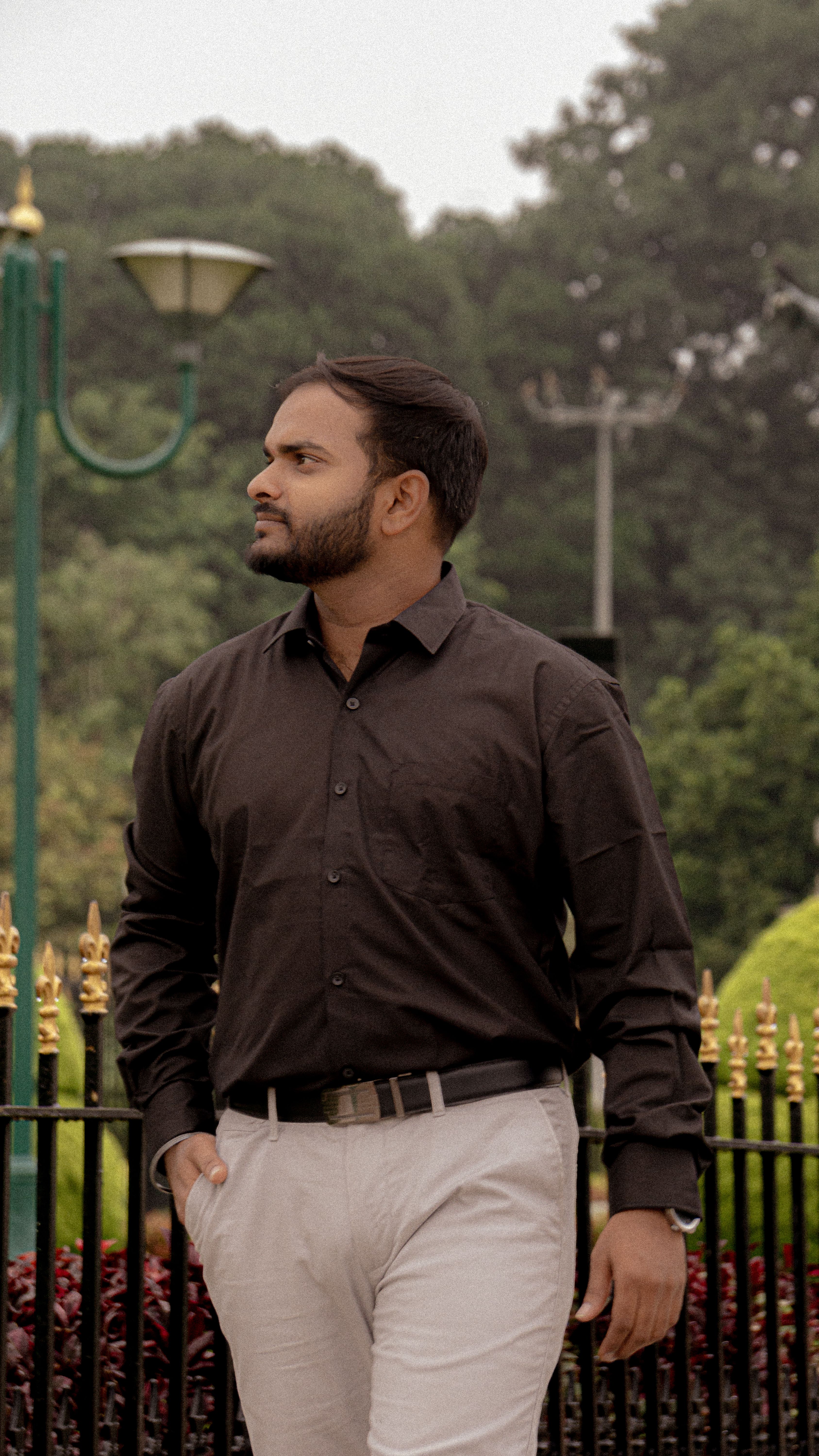 Man in a park with greenery, dressed in a casual shirt and pants.