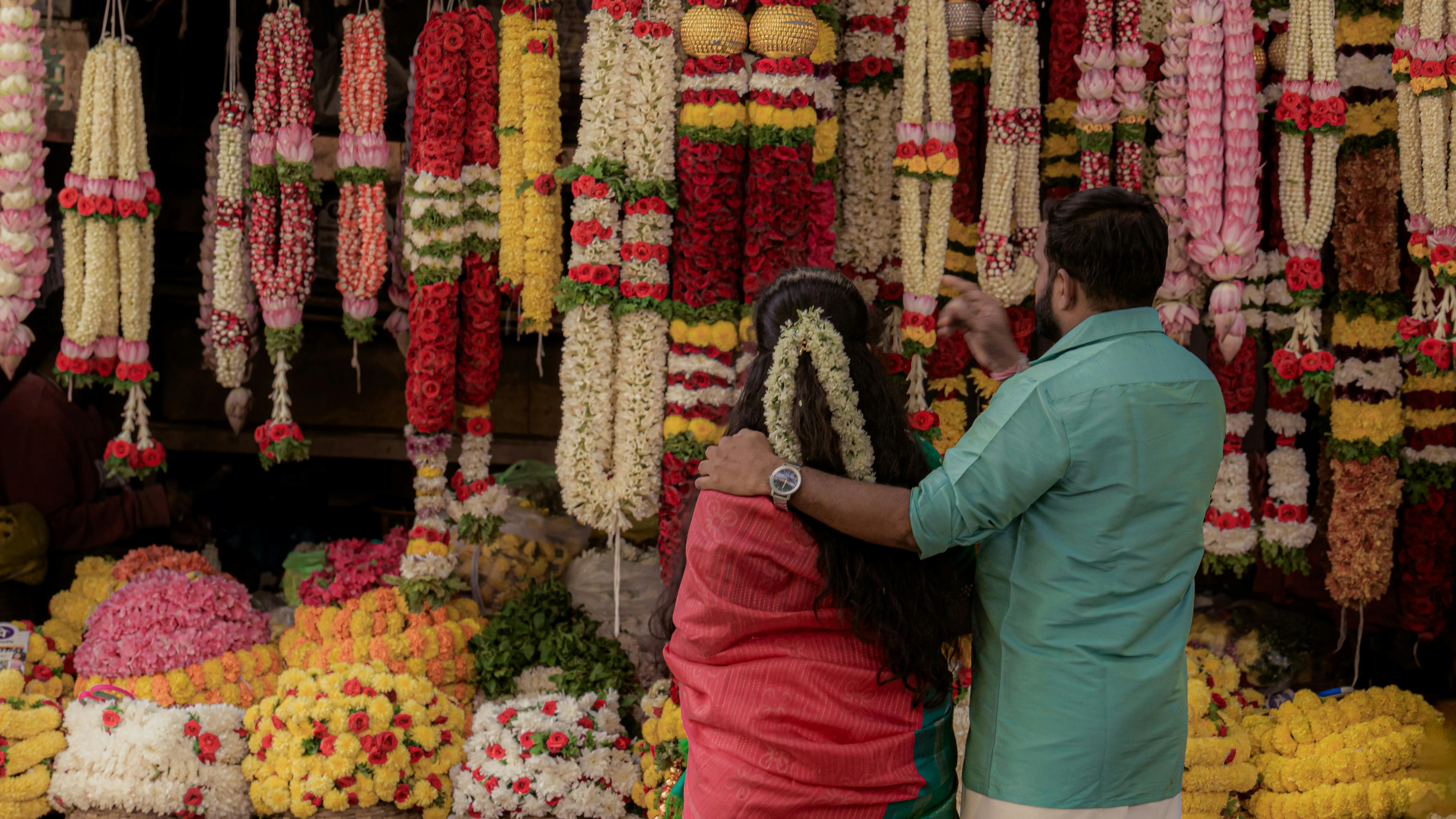 Two people explore a vibrant flower market adorned with intricate garlands.
