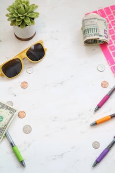 Flat lay of a workspace with cash, coins, pens, sunglasses, and a plant on a marble surface.