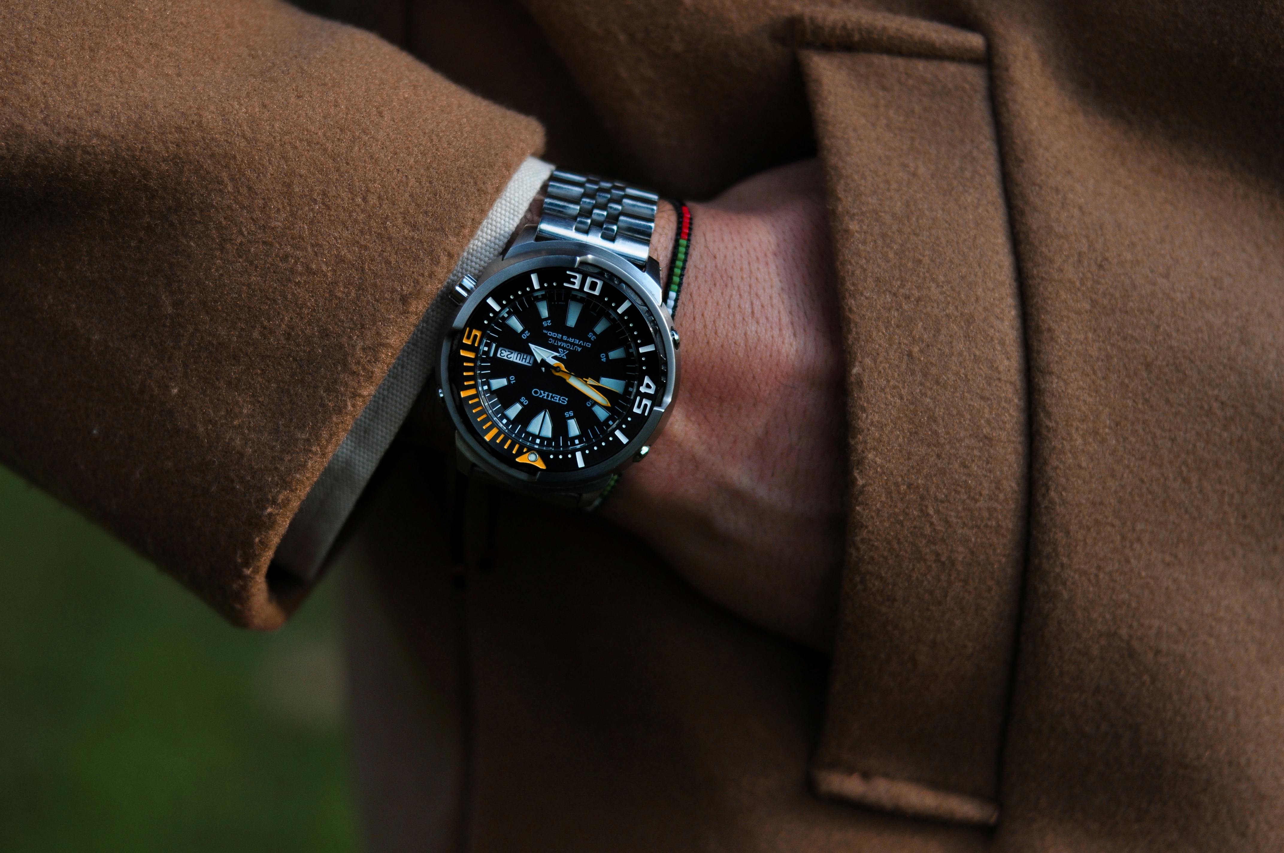 Stylish wristwatch with a metal band on a hand in a brown coat pocket outdoors.