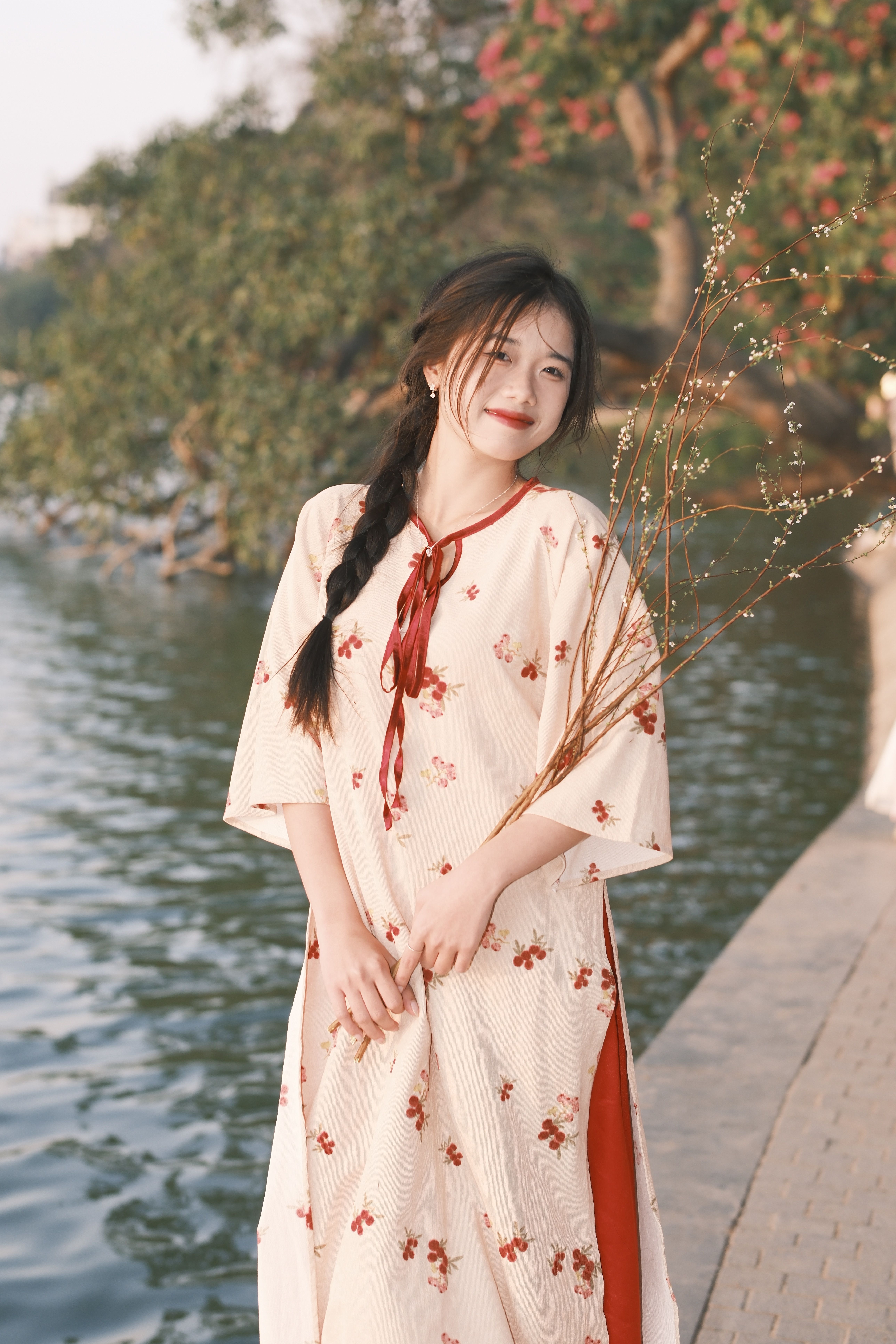 A serene scene of a woman by a lakeside in a floral dress, holding branches.