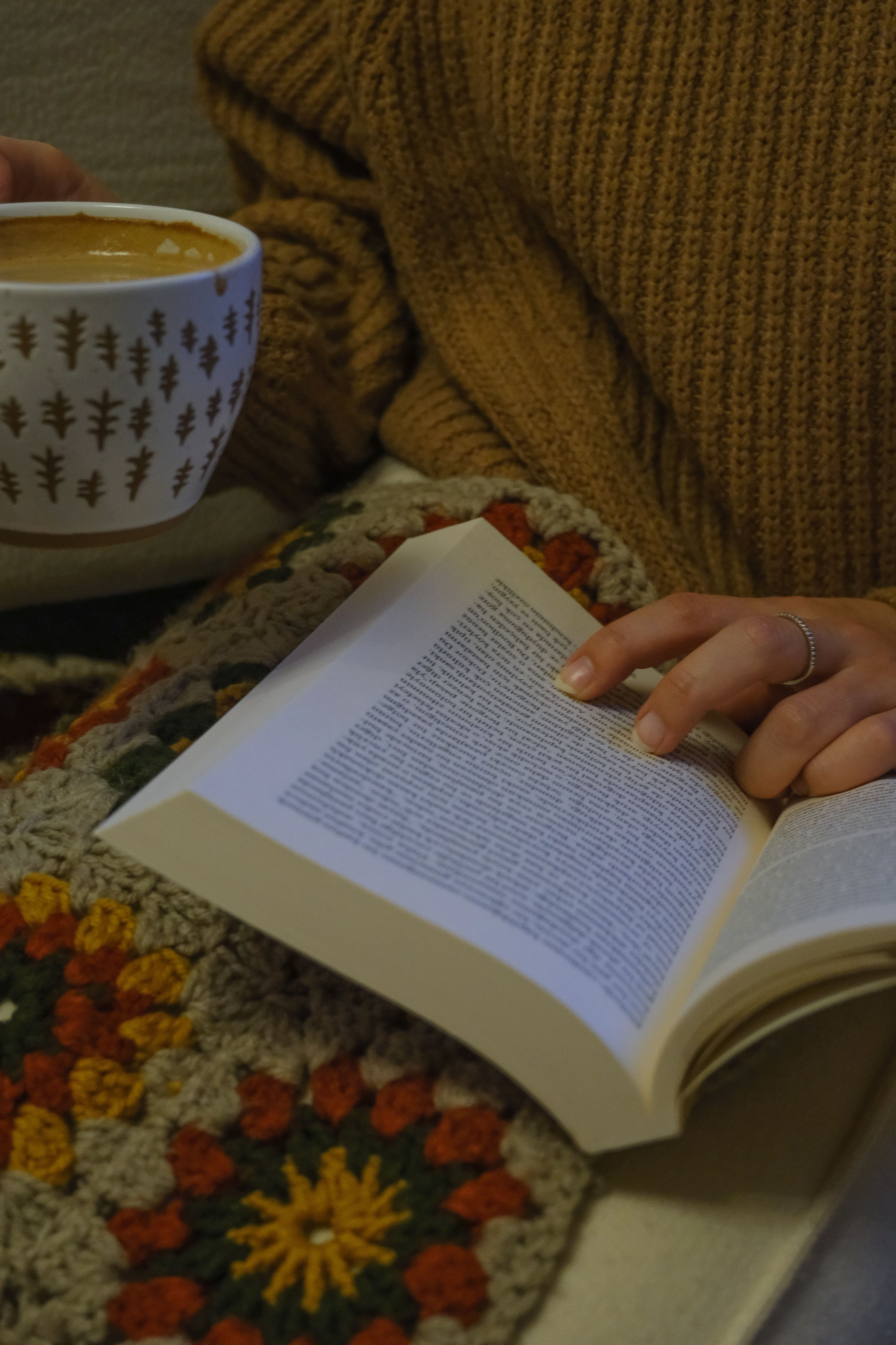 A cozy scene of a person holding a book and coffee mug, wrapped in a colorful blanket.