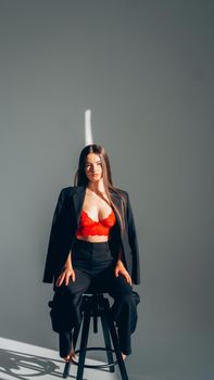 A fashionable woman poses confidently in a black suit and red lingerie, seated on a stool.