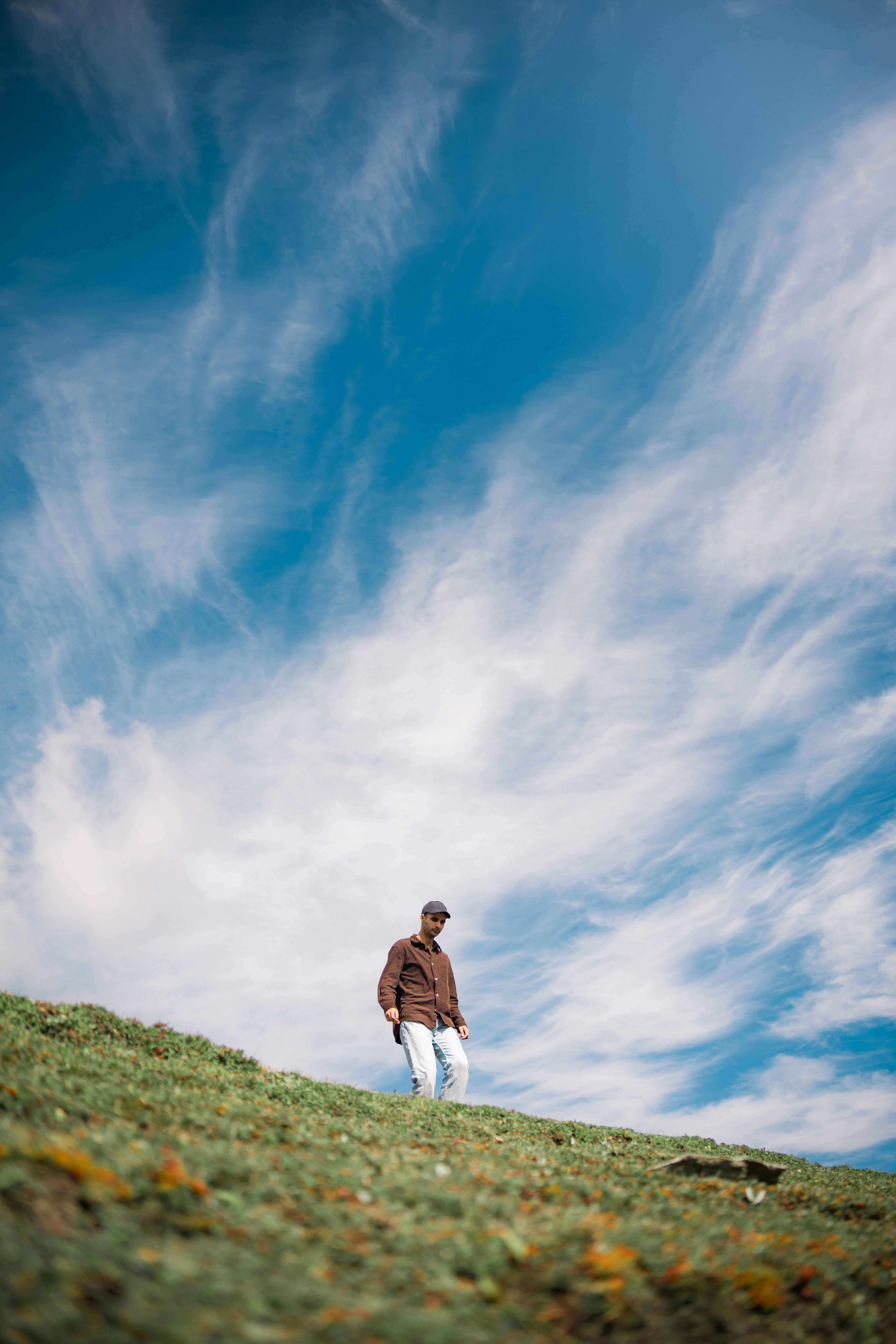 A man in casual attire walks on a grassy hillside against a backdrop of a clear blue sky with scattered clouds.