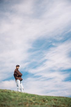 A person stands alone on a grassy hill under a vast, cloudy sky, showcasing solitude and nature.