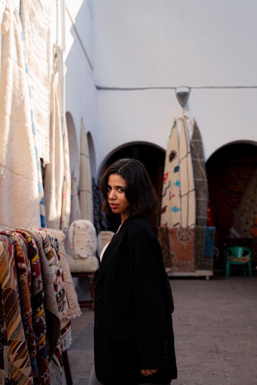 Free A woman in a dark jacket stands in a bazaar with colorful rugs displayed on the walls. Stock Photo