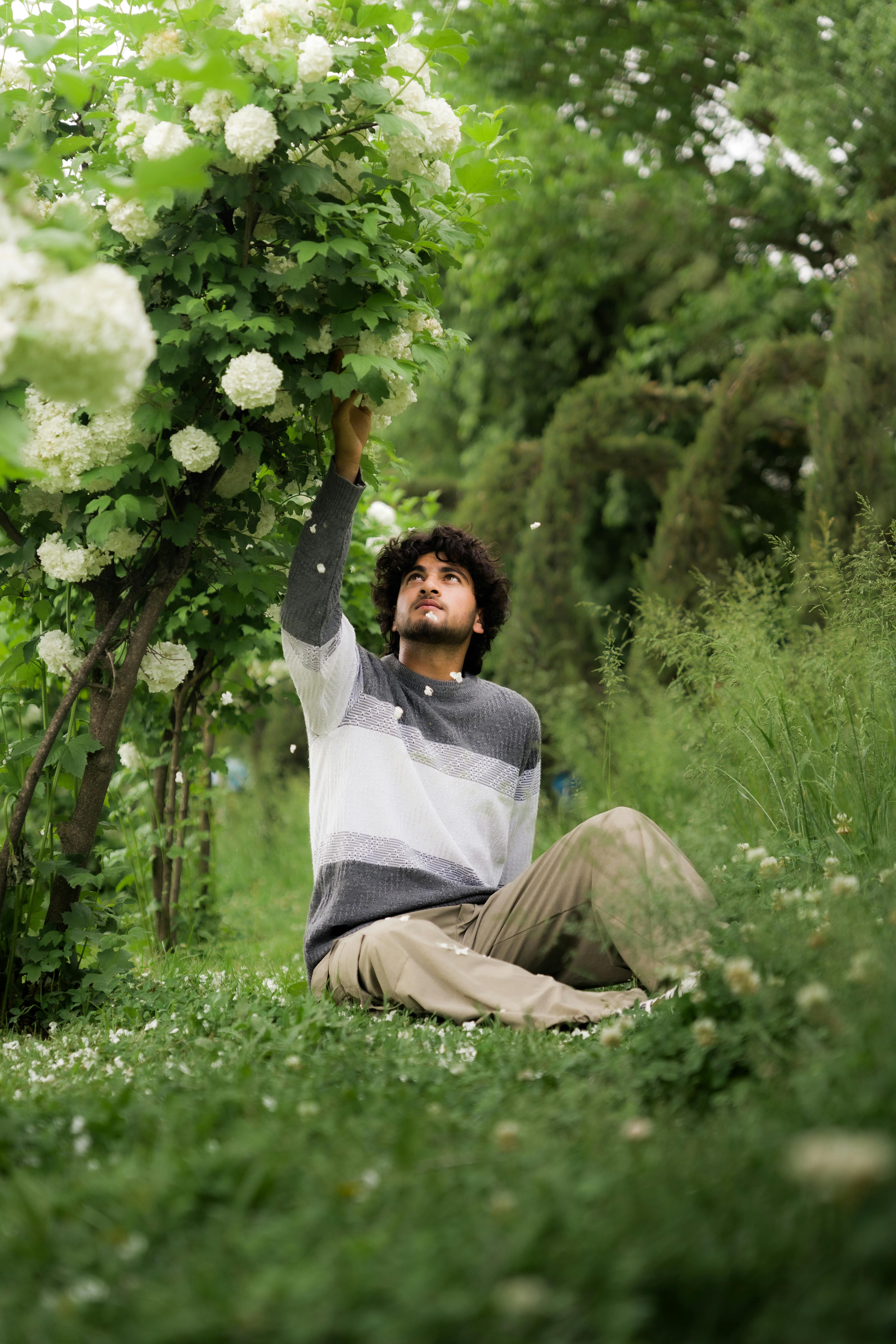 A man in casual attire sits on lush grass, reaching for flowers in a verdant garden.
