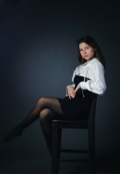 Studio portrait of a woman in a black dress and white blouse sitting on a chair against a dark background.