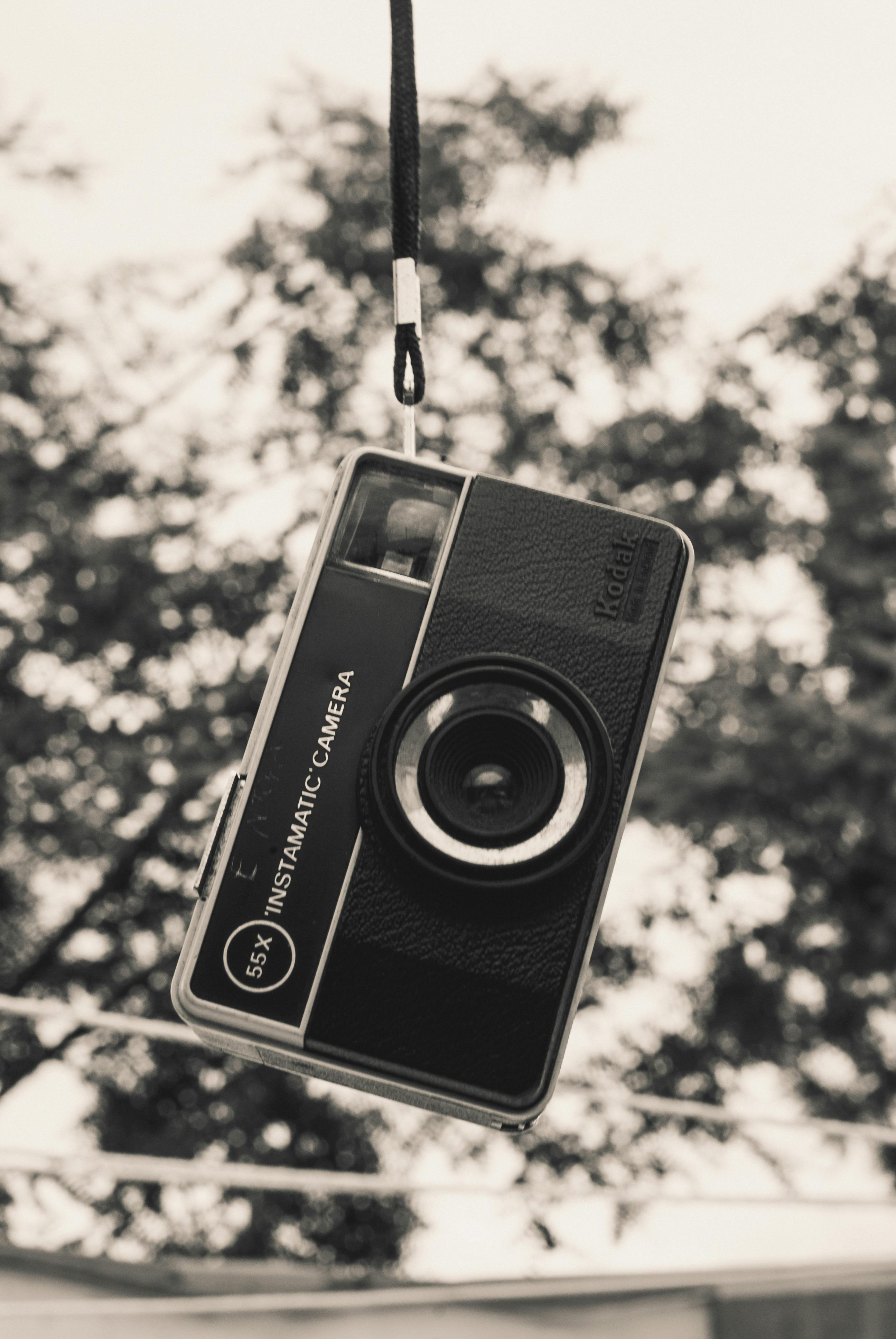 Gratuit Fotografie monocromă a unei camere instantanee clasice suspendate în aer, cu copaci încețoșați în spate. Fotografie de stoc