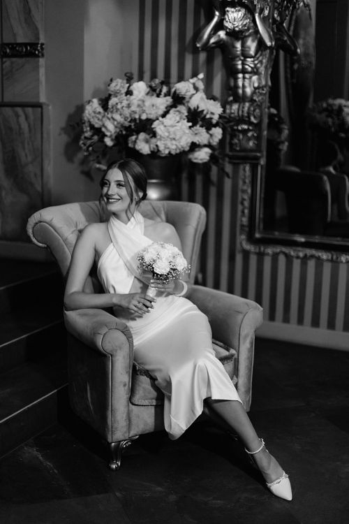 Free A stylish bride in a vintage chair, captured in black and white, holding a bouquet and smiling. Stock Photo