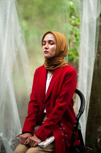 Elegant portrait of a woman wearing a red coat and hijab, sitting outdoors with closed eyes.