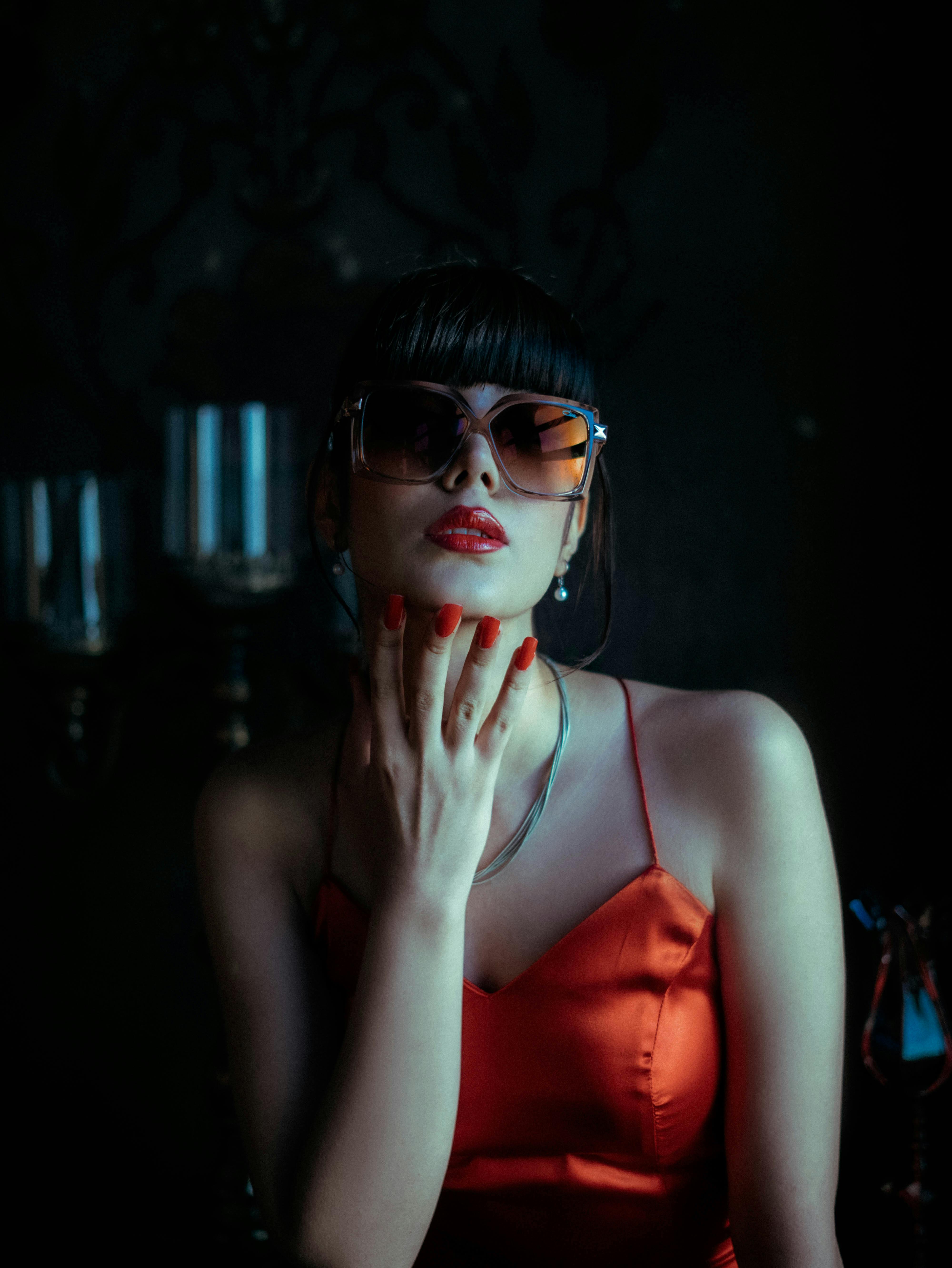 Fashionable woman in sunglasses and red dress posing with elegance under moody lighting.