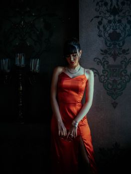 Elegant woman in a red dress stands in a dimly lit room next to vintage decor, creating a moody atmosphere.