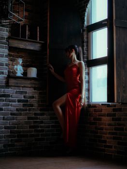 A woman in a red dress leans against a brick wall in a dimly lit room, exuding elegance.