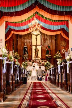 Bride and groom at altar in colorful, ornate church with floral decorations.