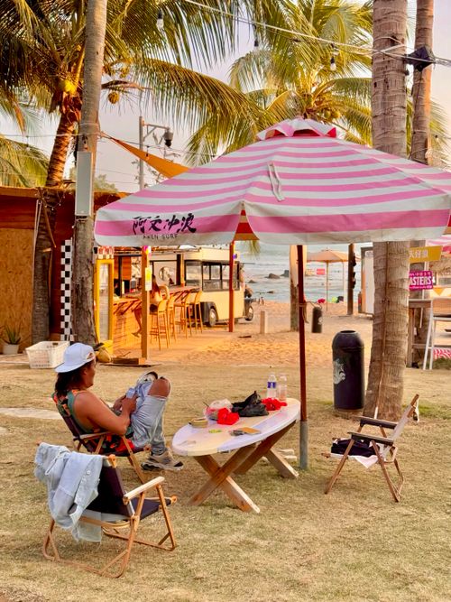 Free A person relaxes at a beachside cafe under a pink striped umbrella, with palm trees and a serene ocean view. Stock Photo