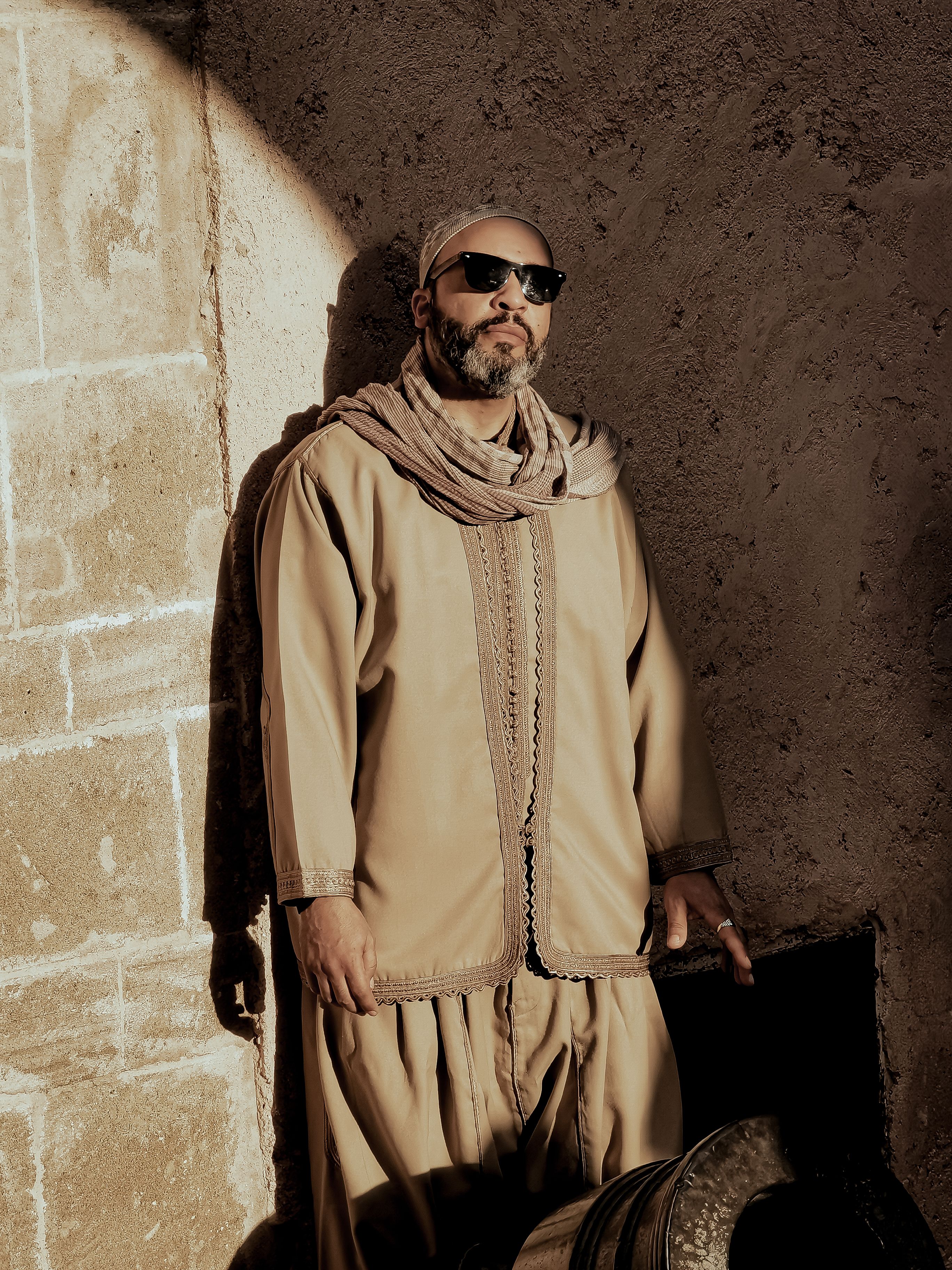 Man in traditional Moroccan attire with sunglasses, posing against a textured wall.