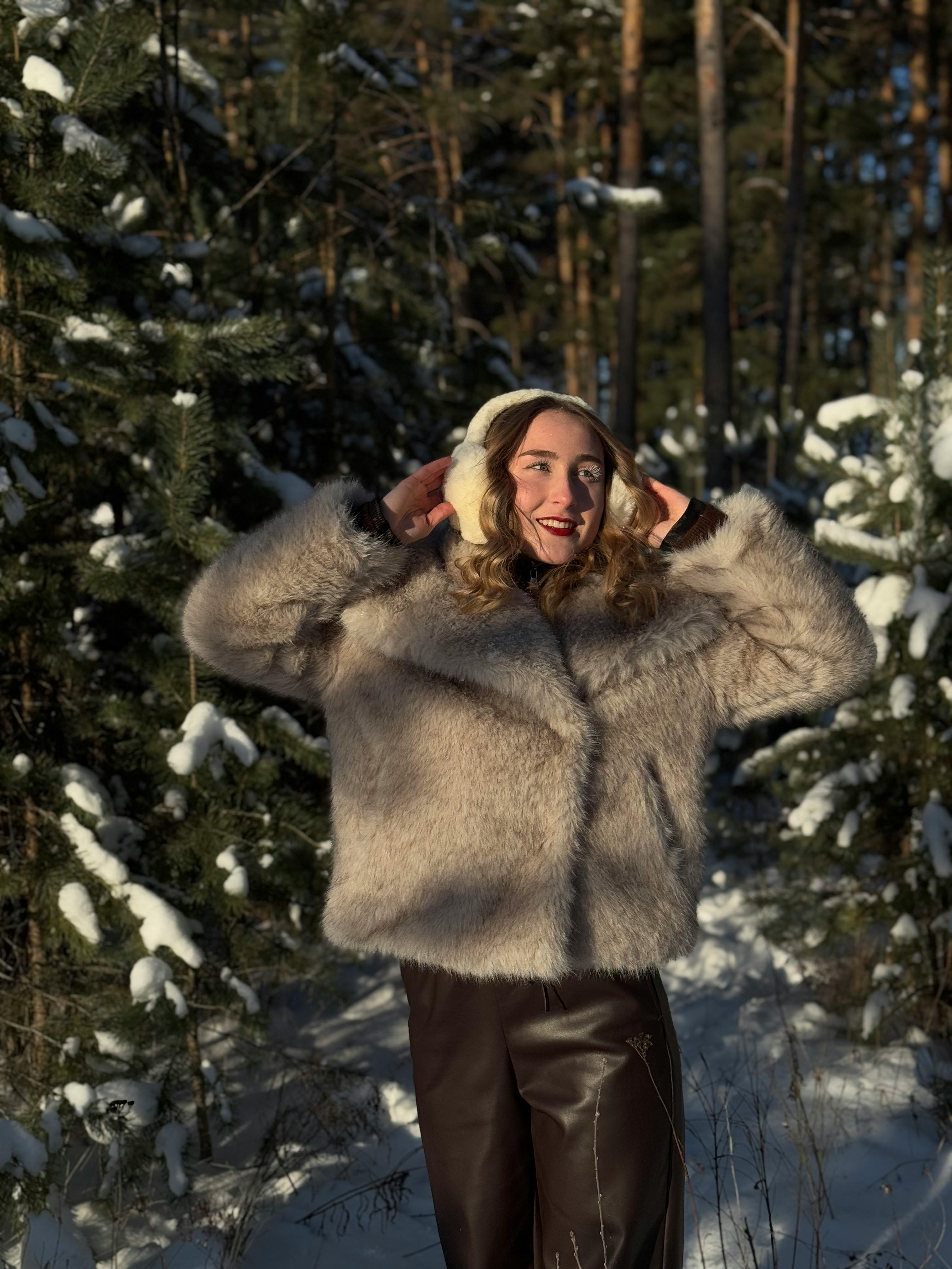 Smiling woman in fur coat and earmuffs standing in a snowy forest setting.