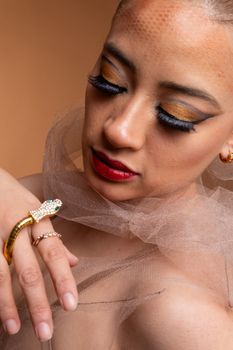 Close-up portrait of a woman with golden snake jewelry and artistic makeup against a mocha backdrop.