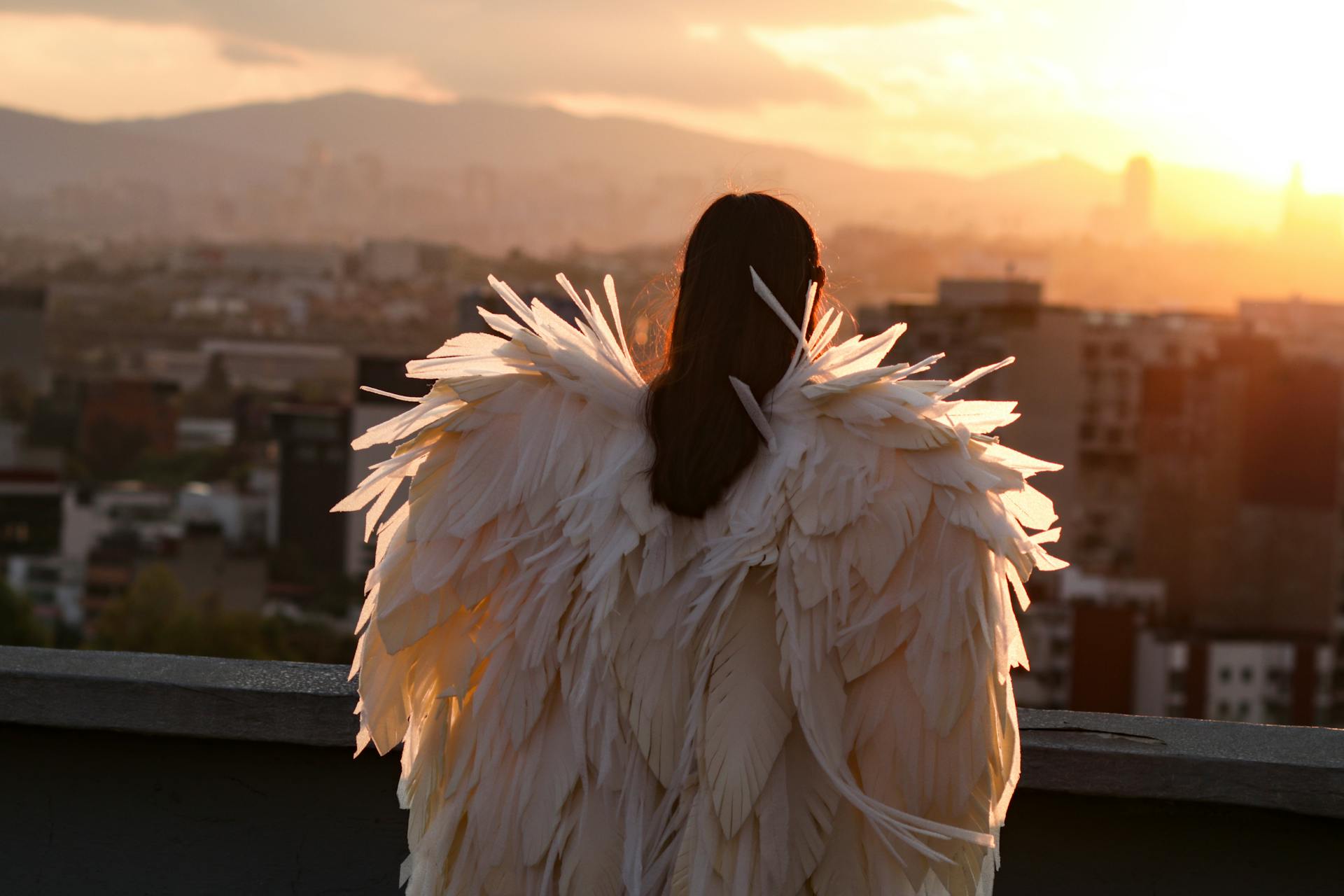 A woman in an angel costume gazes over a cityscape as the sun sets, casting a glowing light.