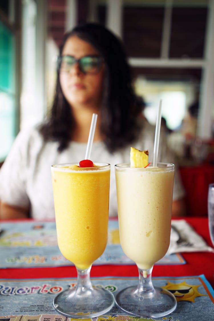 Woman Wearing White Crew-neck T-shirt And Eyeglasses Sitting Near Table And Two Juice In Glasses