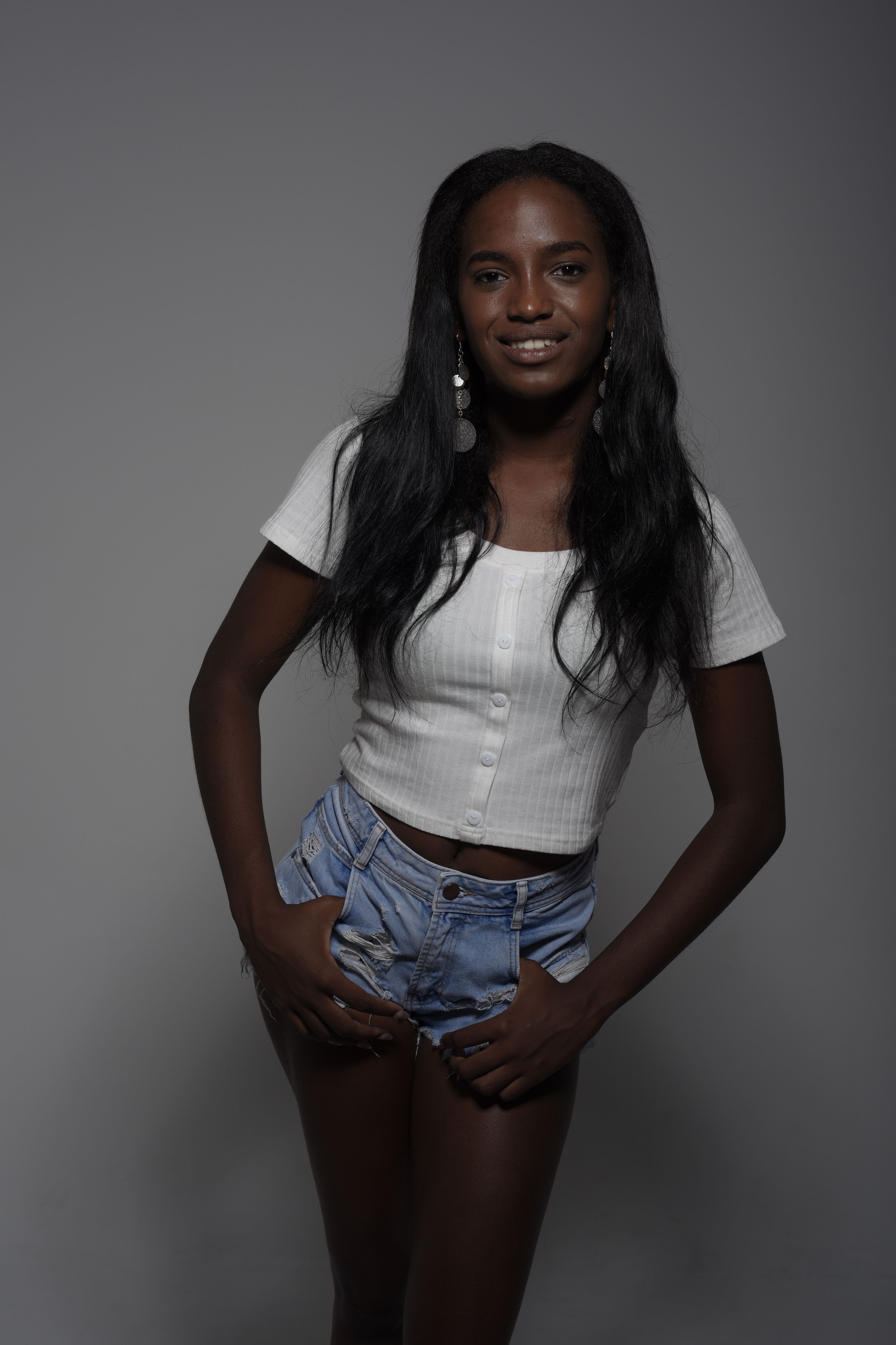 A confident woman poses in a white top and denim shorts with a neutral background.