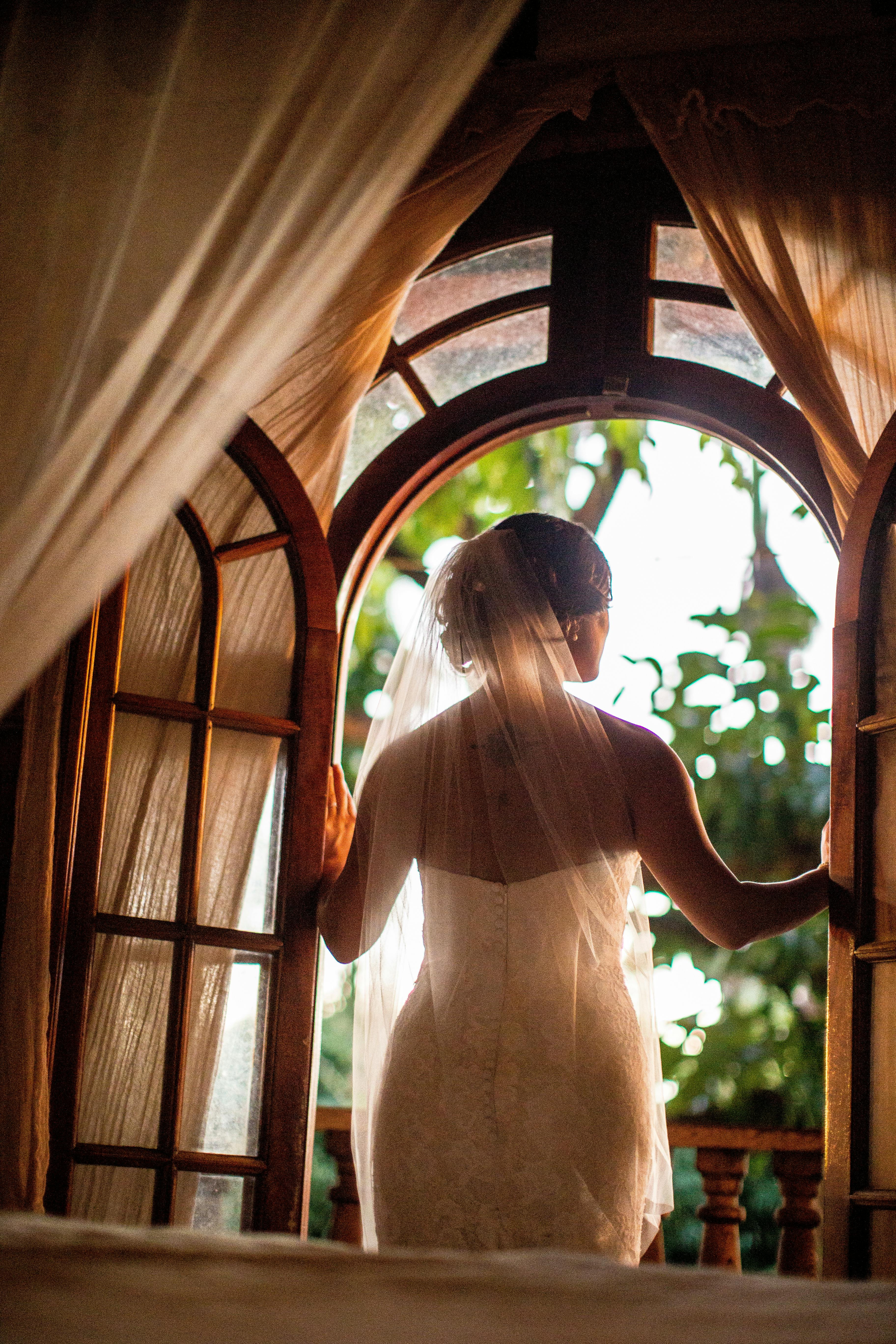 Bride in wedding gown standing by arched window, captured from behind during sunset.