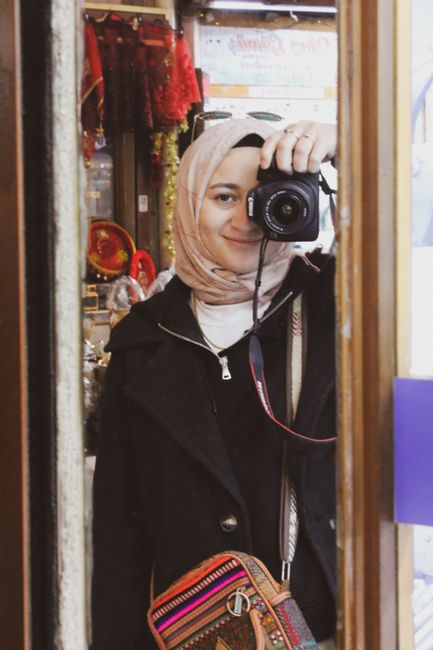 A woman in a hijab takes a mirror selfie inside a store, showcasing her camera and colorful bag.