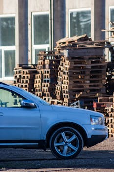 Photo of a car beside a pile of wooden pallets at an industrial site.