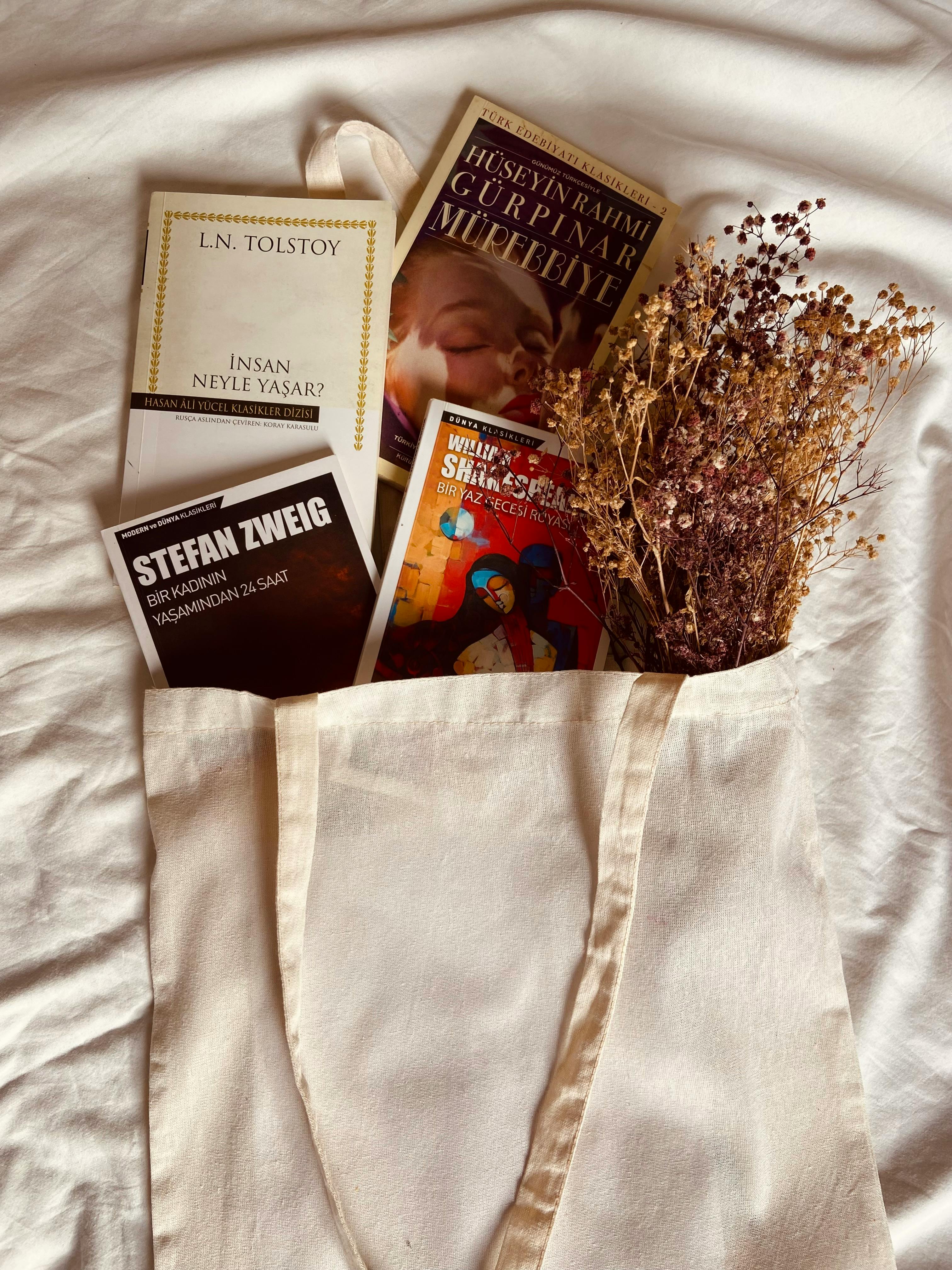 Beige tote bag filled with classic books and dried flowers on a soft white cloth.