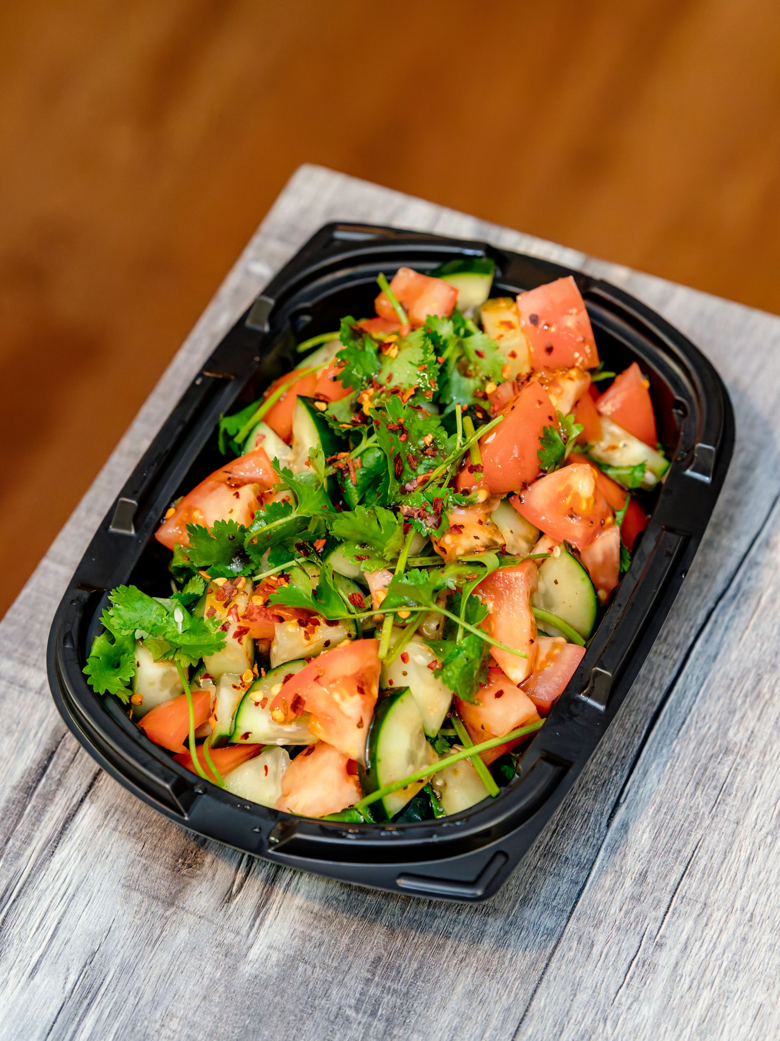 Vibrant tomato and cucumber salad with coriander leaves in a black tray.