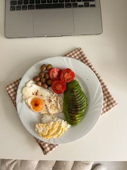 Delicious breakfast of eggs, avocado, tomatoes, and olives by laptop.