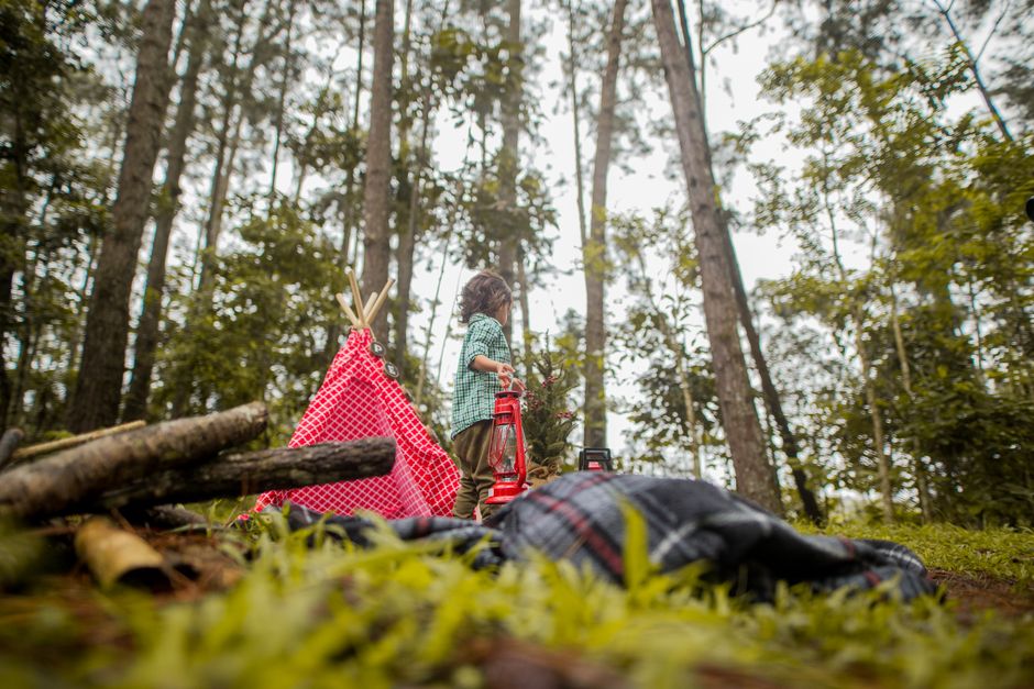 A young child explores a scenic forest campsite with a tent and lantern, surrounded by nature.