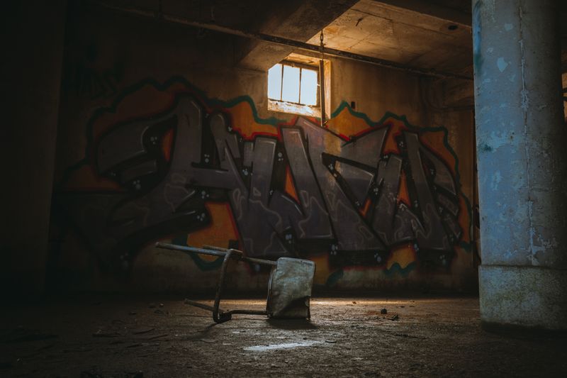 Dimly lit abandoned building interior with a large colorful graffiti piece on the wall, light streaming through a window