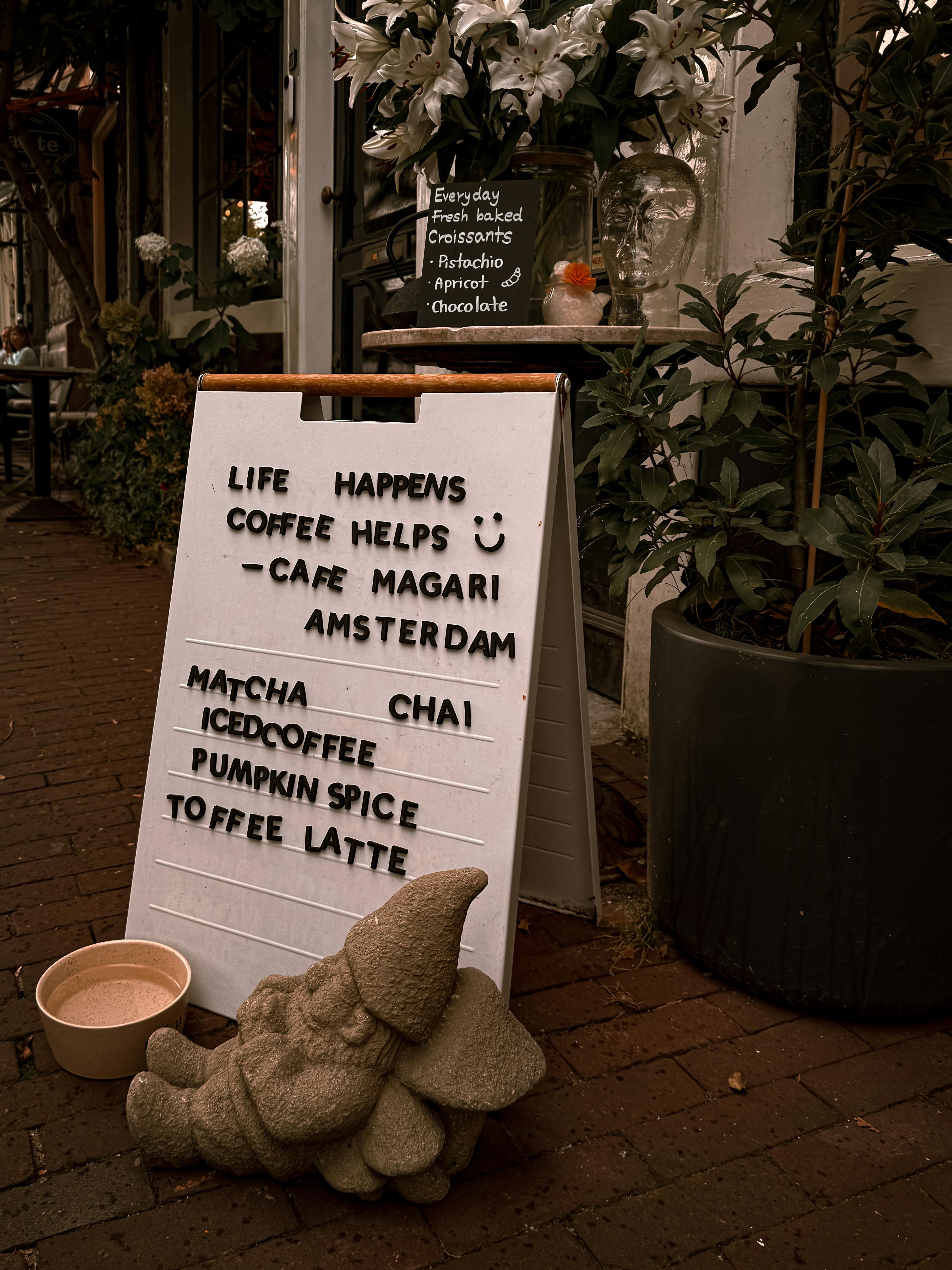 A cozy café entrance in Amsterdam featuring a playful coffee sign and gnome statue outdoors.