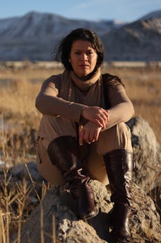 Woman in boots sitting on rock with scenic mountain backdrop at daytime.