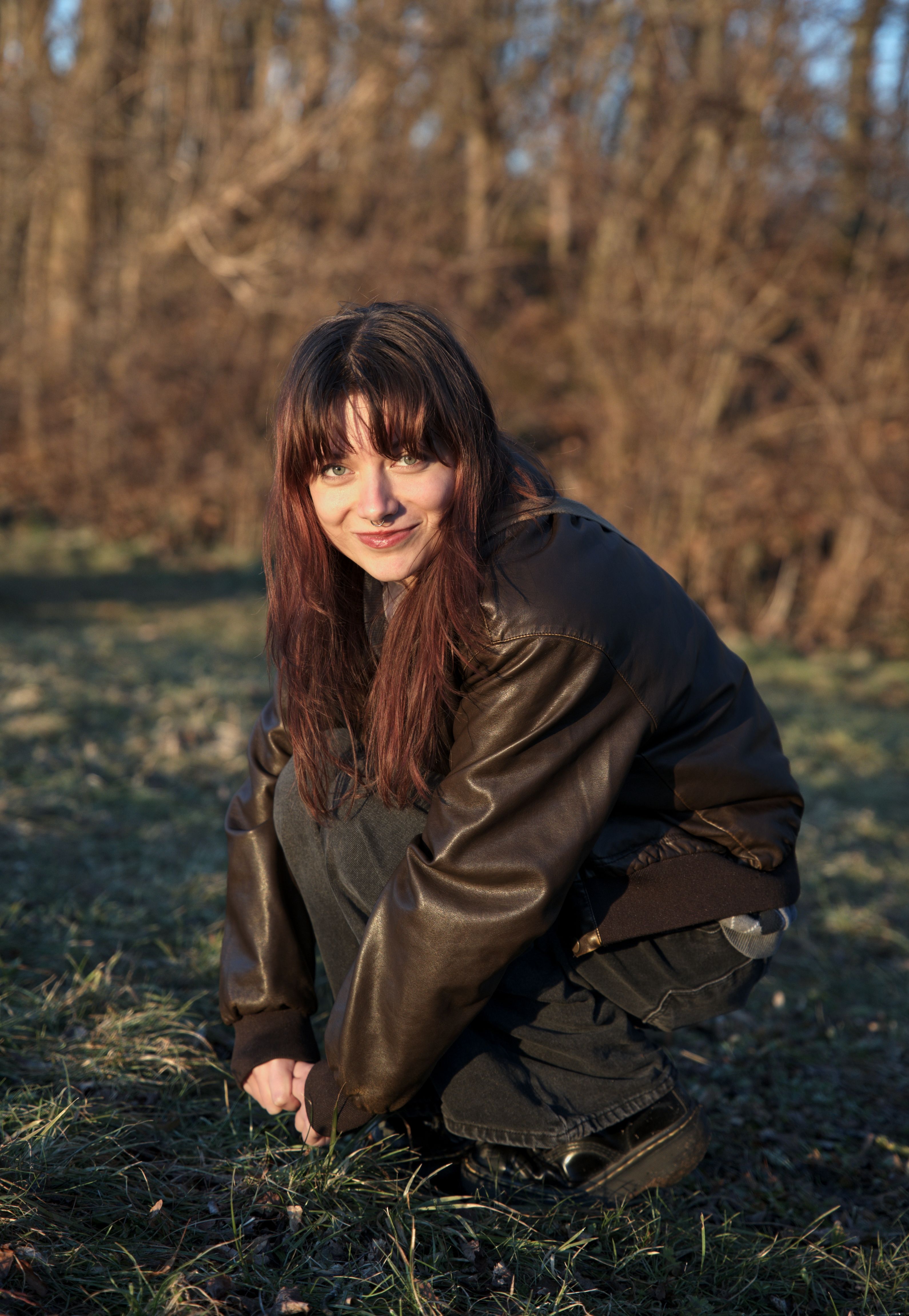 Smiling young woman crouching in a forest wearing a leather jacket, enjoying the sunlight.