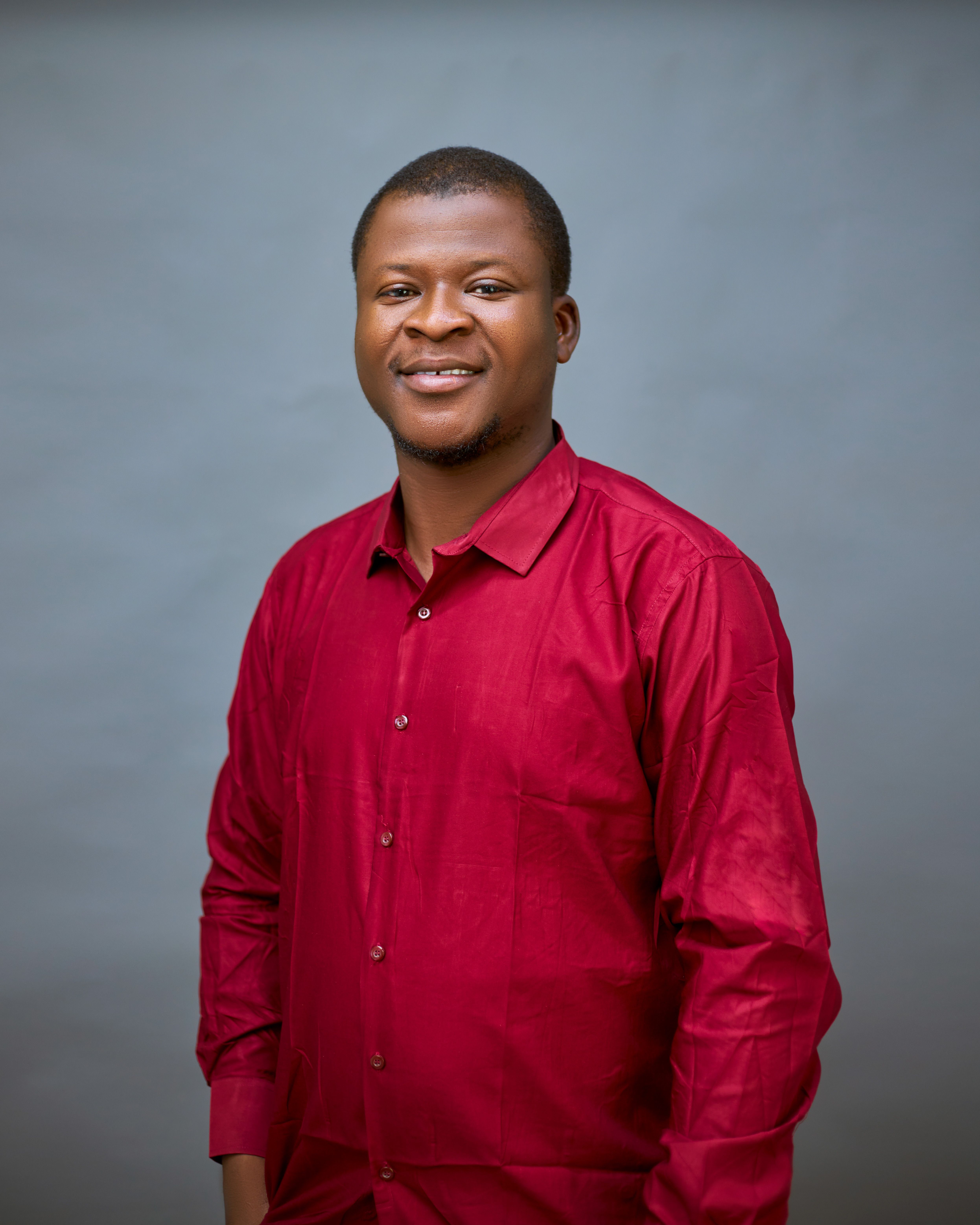 Portrait of a smiling African adult male in a red shirt against a plain background.