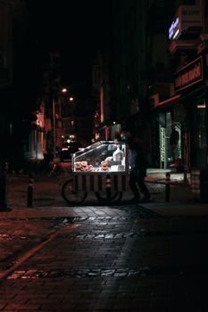 A street vendor sells food from an illuminated cart at night in a dimly lit urban area.