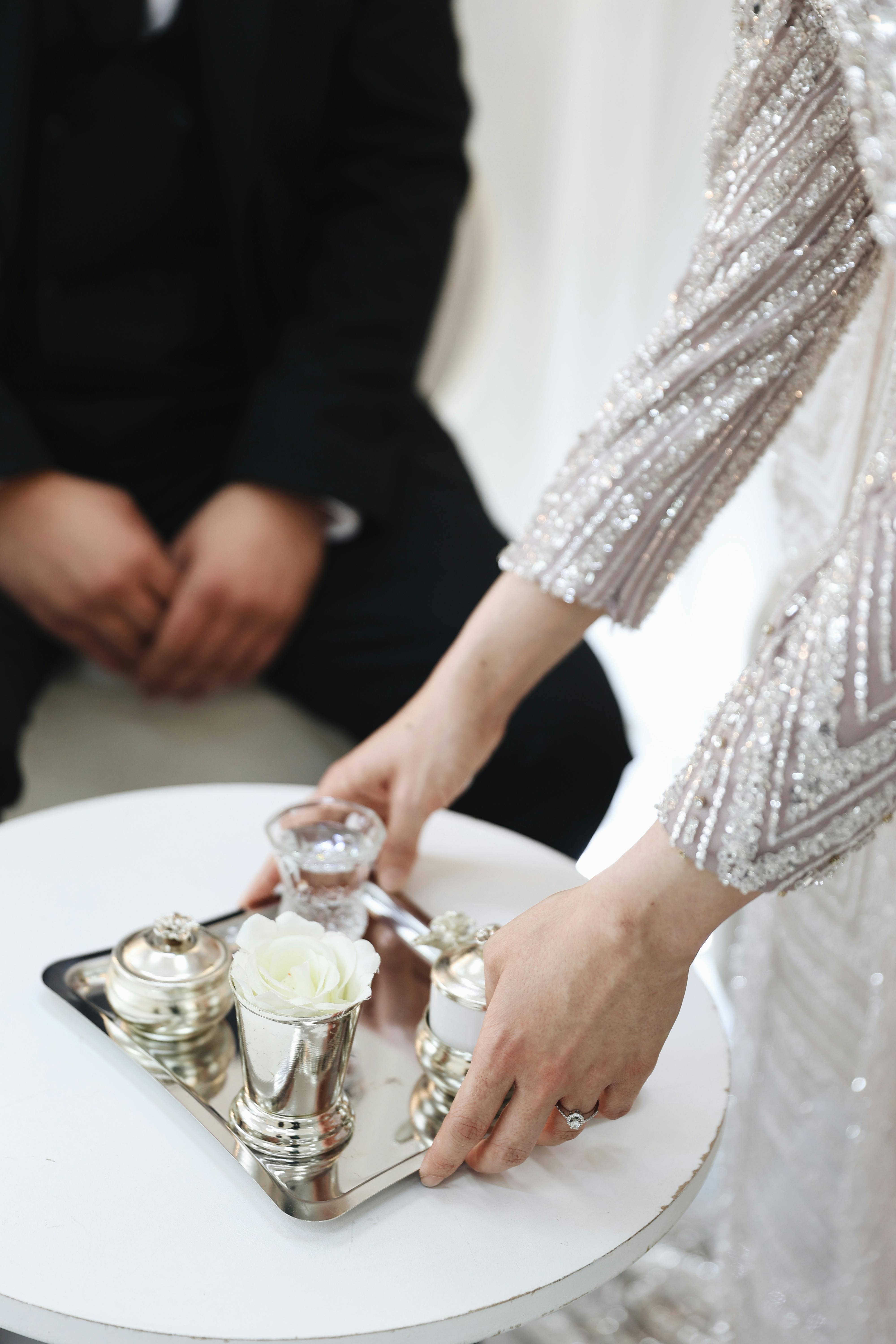 A person in a formal gown prepares a tea service on a small table, featuring silverware and a white rose.
