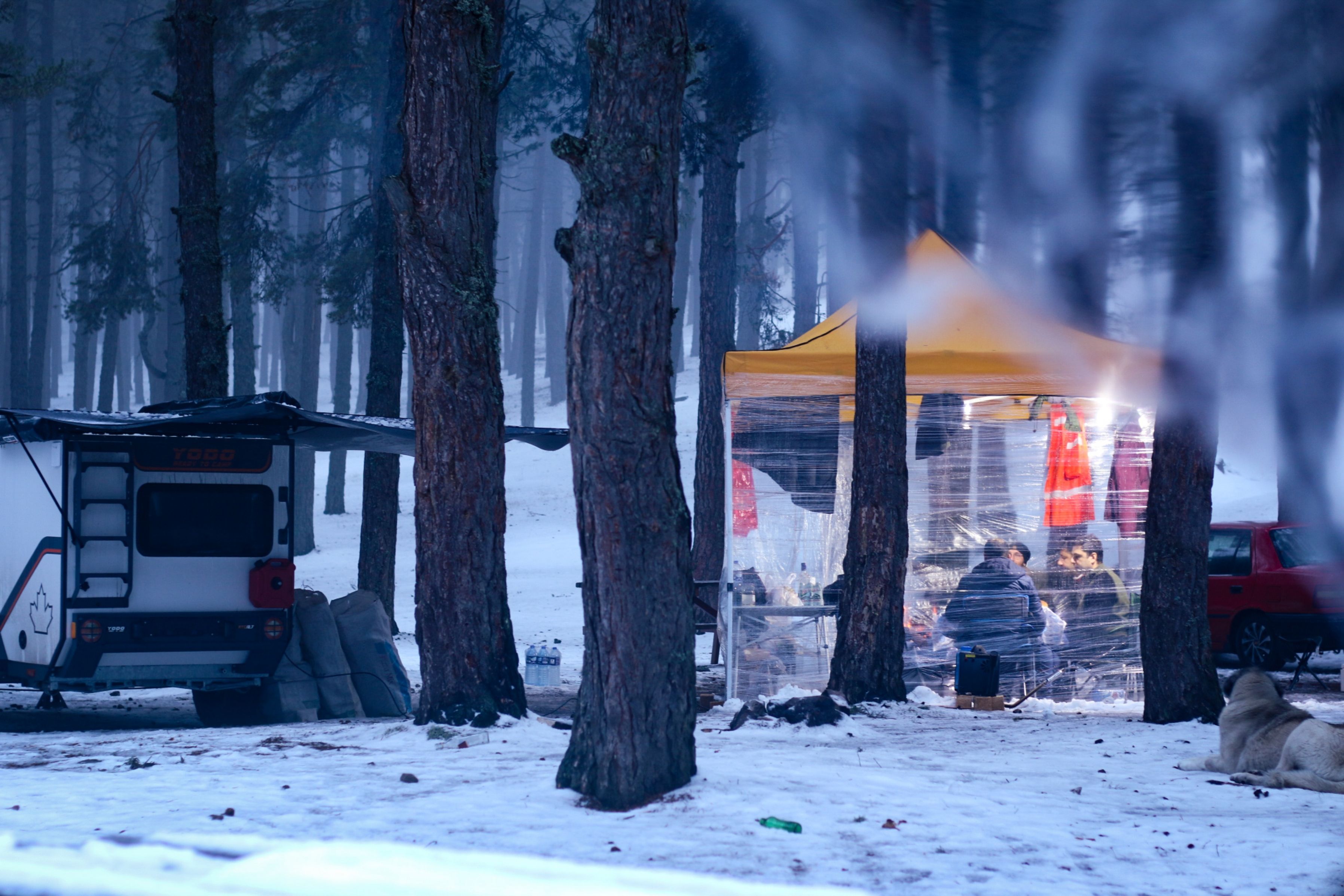 A cozy winter campsite with a tent and camper in a snowy forest setting.